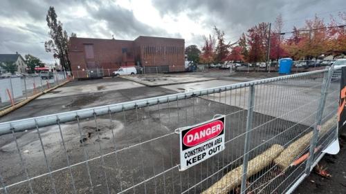A wet and empty lot surrounded by a construction fence on a rainy day with dark grey clouds in the sky. A sign on the fence says "Danger, Construction Area, Keep Out."