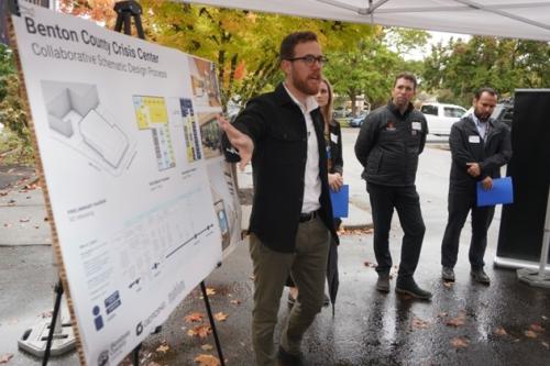 Man wearing glasses and a suit stands under a canopy next to three others on a rainy day and gestures to an architectural drawing on an easel.