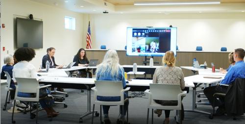 People sitting around a conference table hold a meeting with virtual attendees on a large TV in the background.