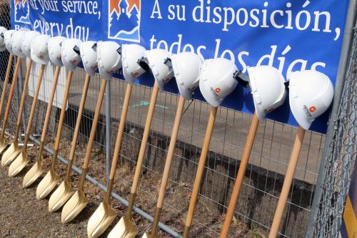 A row of golden shovels topped with hard hats lean against a blue sign hanging on a construction fence.