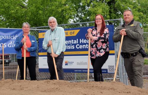 Three people and a uniformed law enforcement officer stand near a pile of dirt with shovels in hand.