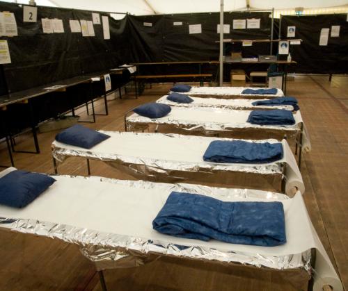 Five cots each lined with exam room paper and topped with a blue pillow and blanket are lined up in a row in a large white emergency tent filled with temporary room dividers.