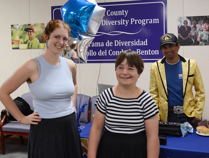 Two people pose and smile in front of balloons and another person wearing a sequined gold jacket.