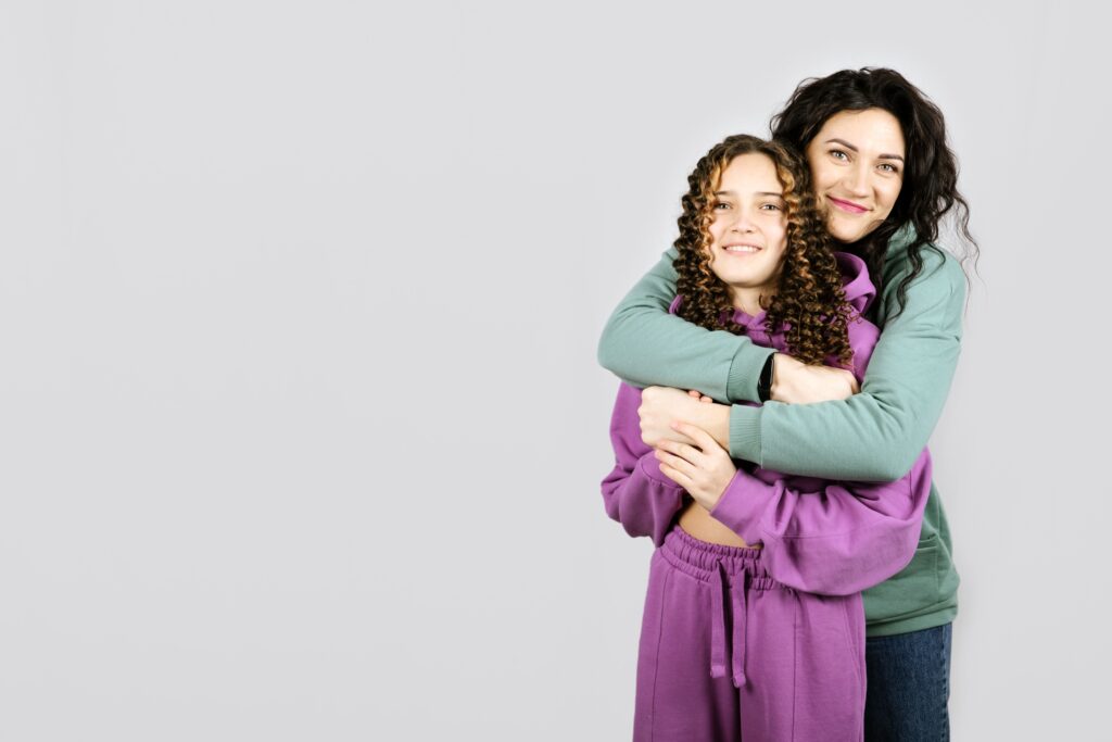 A woman in a teal sweatshirt and jeans hugs her teenage daughter wearing a purple sweat suit.