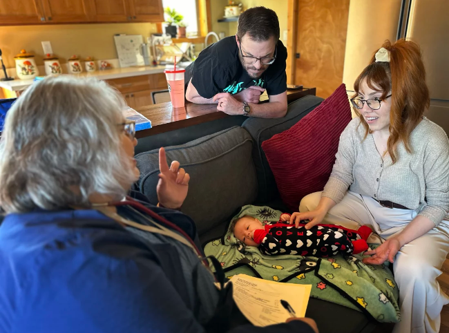 A nurse wearing a blue smock talks with a family on their living room couch.