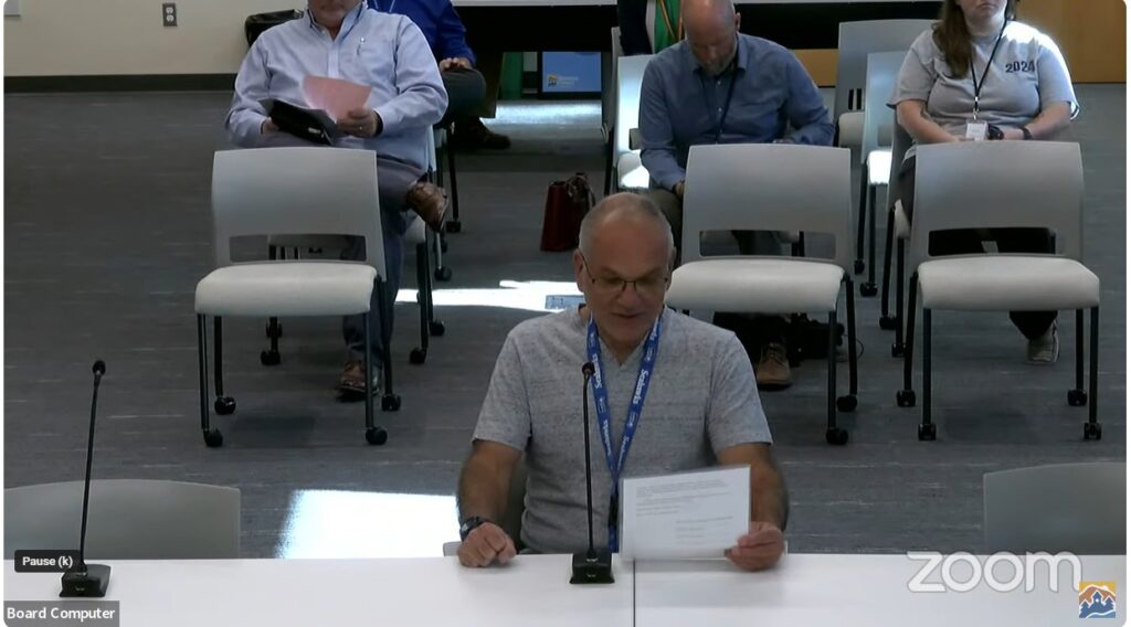 A man sitting at desk speaking into a microphone and holding a paper.