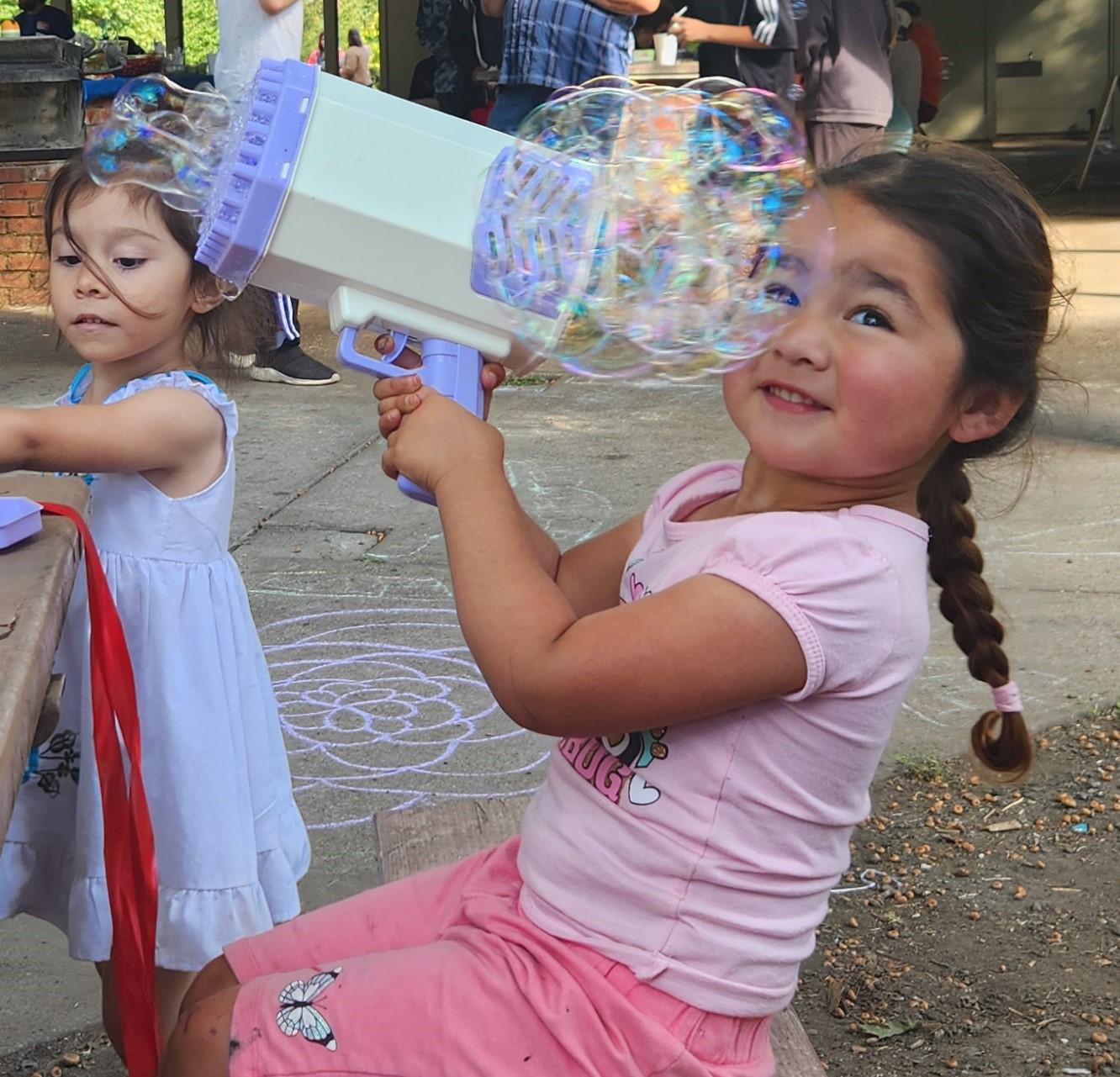 A young child wearing a pink outfit with braided hair plays with a bubble blower.