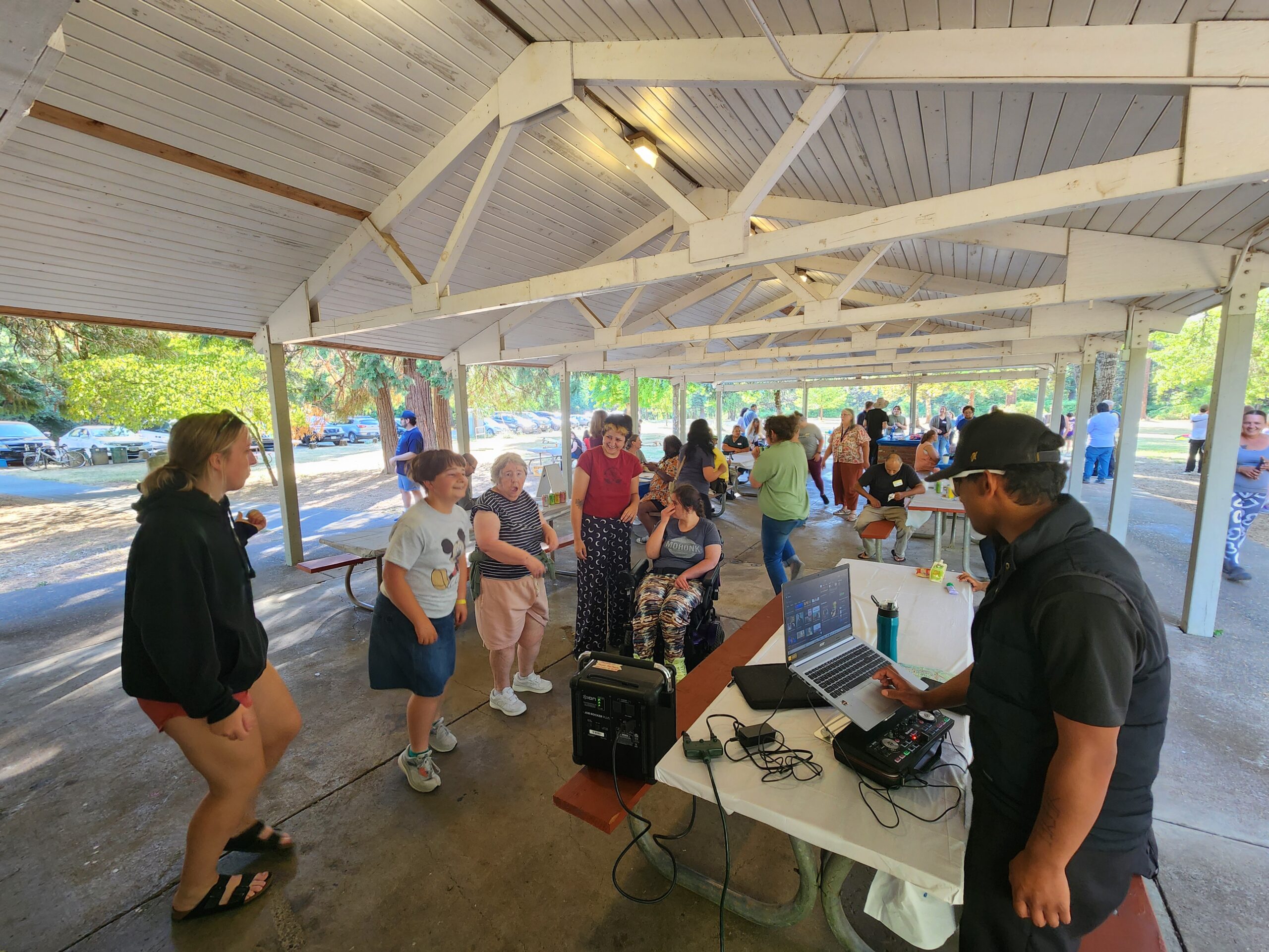 A DJ plays music for a crowd of dancing people in a covered area in a park.