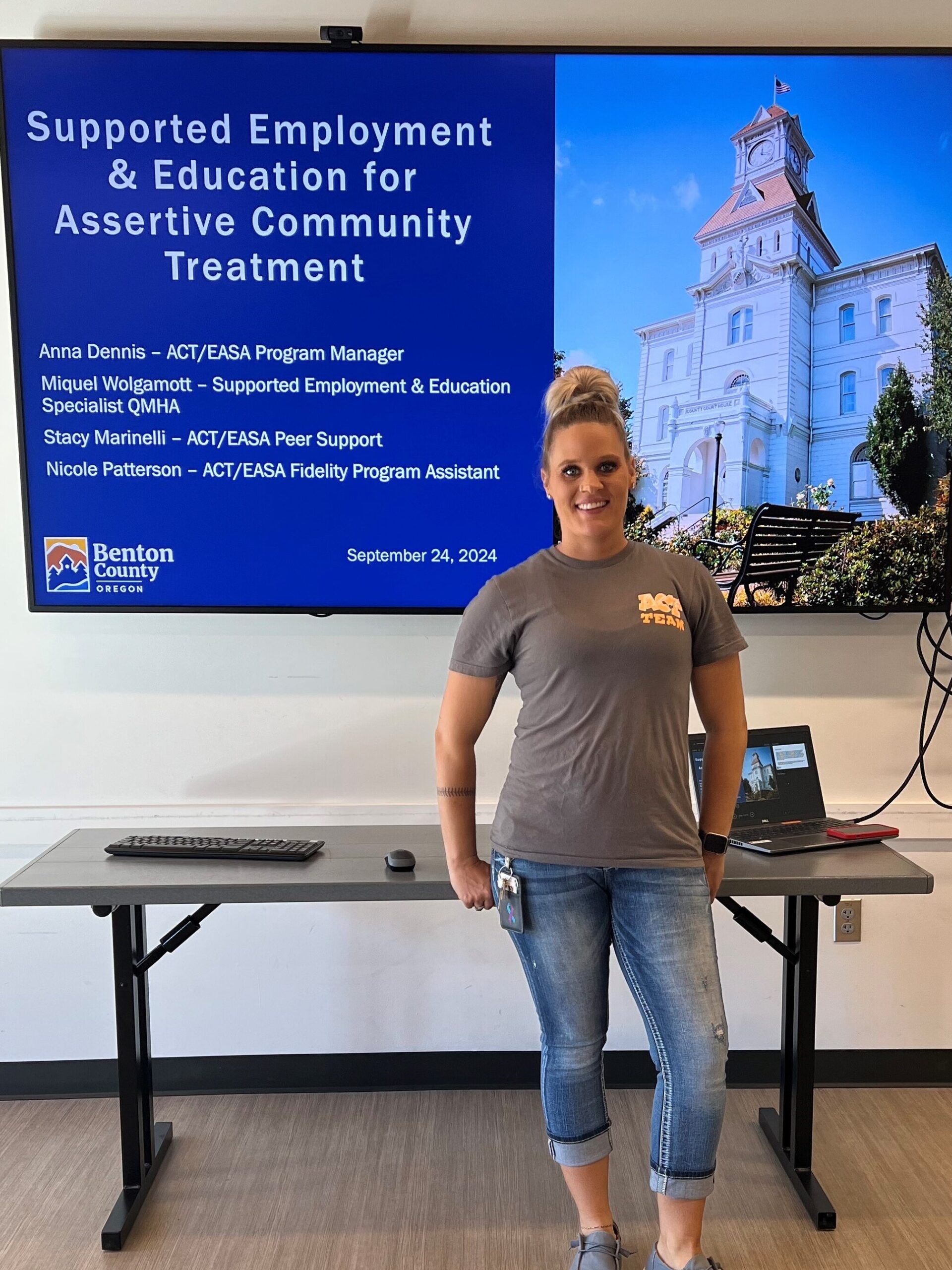 A person poses next to a monitor displaying a PowerPoint presentation.