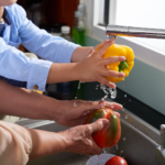 A child and an adult wash yellow and red bell peppers under a kitchen faucet.