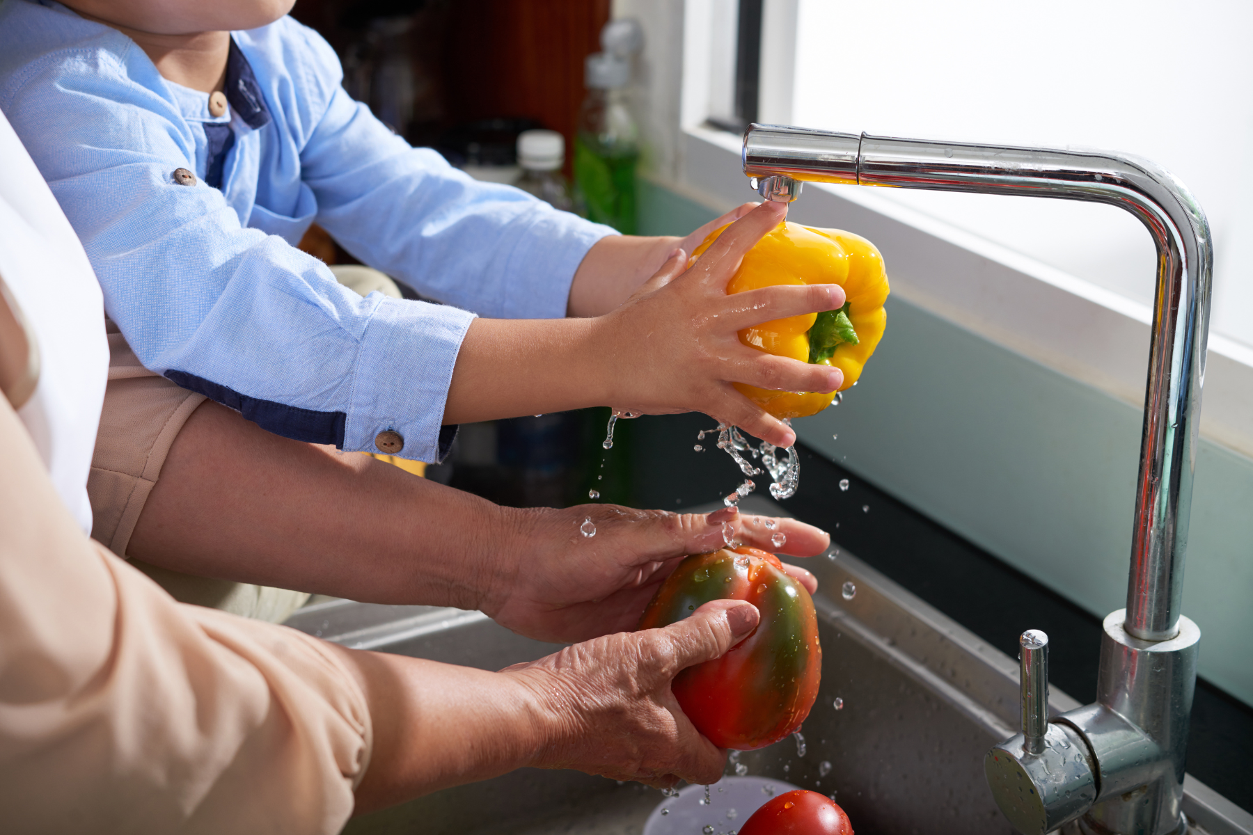 A child and an adult wash yellow and red bell peppers under a kitchen faucet.