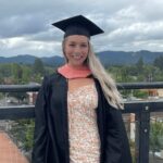 A woman with blonde hair wearing a lack graduation cap & gown with an orange hood.