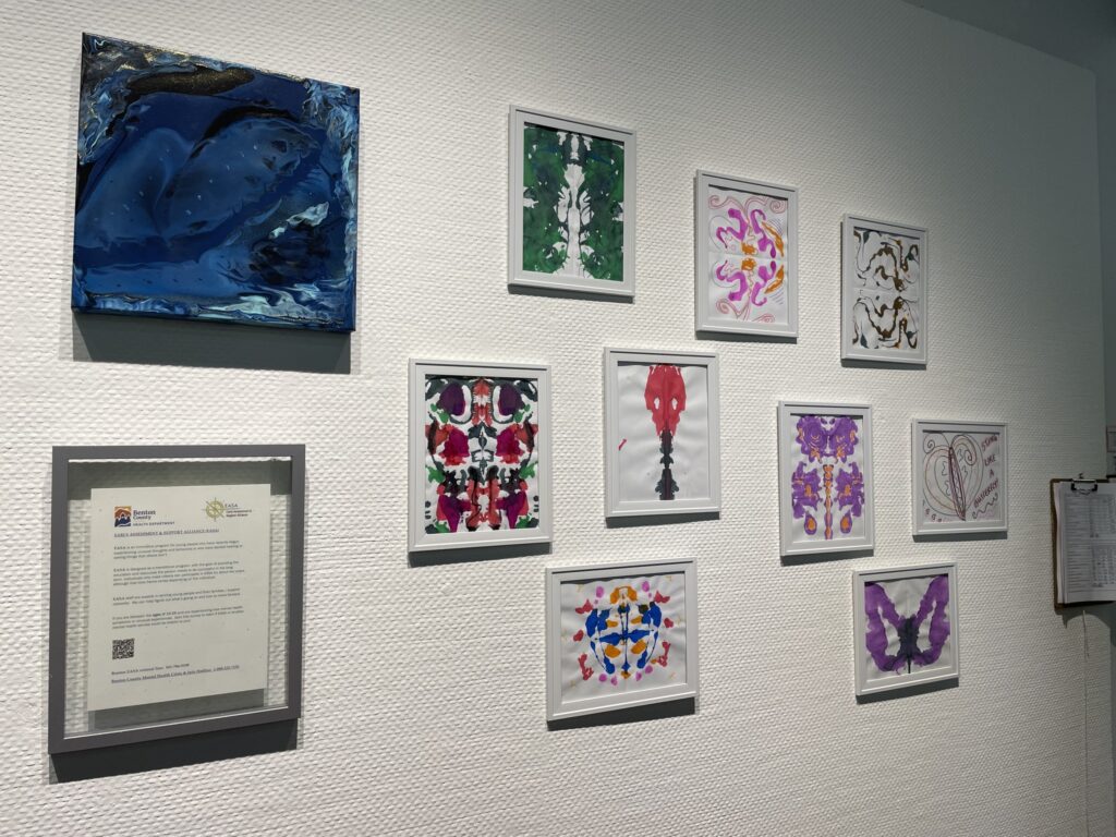 Various painted artworks hang on a white wall. Some works contain butterfly-like imagery in shades of purples, reds, pinks, oranges, blues, and greens. Some art is more abstract.