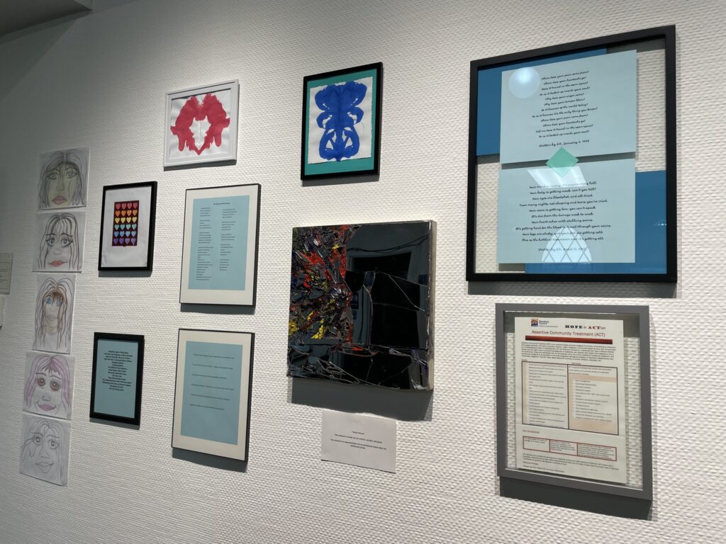 Various framed artworks hang on a white wall. Some works are hand drawn portraits of female faces. Some are more abstract.