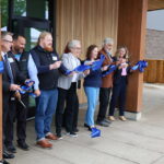 Eight smiling people hold scissors while cutting a blue ribbon tied between two posts at the entrance of a building.