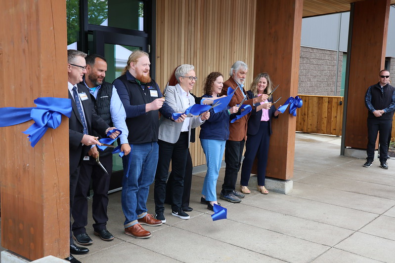 Eight smiling people hold scissors while cutting a blue ribbon tied between two posts at the entrance of a building.