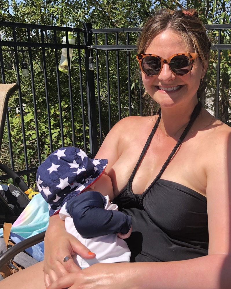 A woman wearing a black swimsuit and sunglasses breastfeeds her baby. /