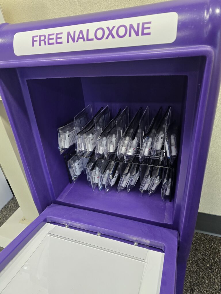 Purple dispenser labeled 'FREE NALOXONE' stocked with multiple naloxone kits in clear plastic packaging.