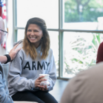 A young woman wearing an Army sweatshirt holds a cup while an older man places a comforting hand on her shoulder during a conversation indoors.