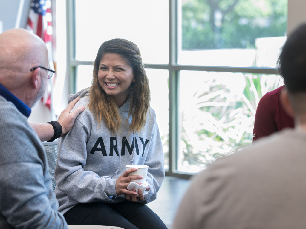 A young woman wearing an Army sweatshirt holds a cup while an older man places a comforting hand on her shoulder during a conversation indoors.