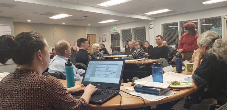 A large group sits around tables in a meeting room, with one person standing and speaking while others listen and work on laptops and papers.