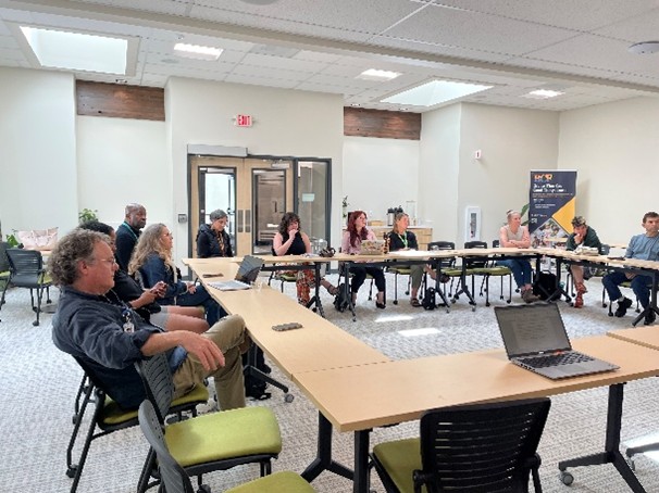 A group of people sit around U‑shaped tables in a bright meeting room, participating in a discussion with laptops and notebooks on the tables.