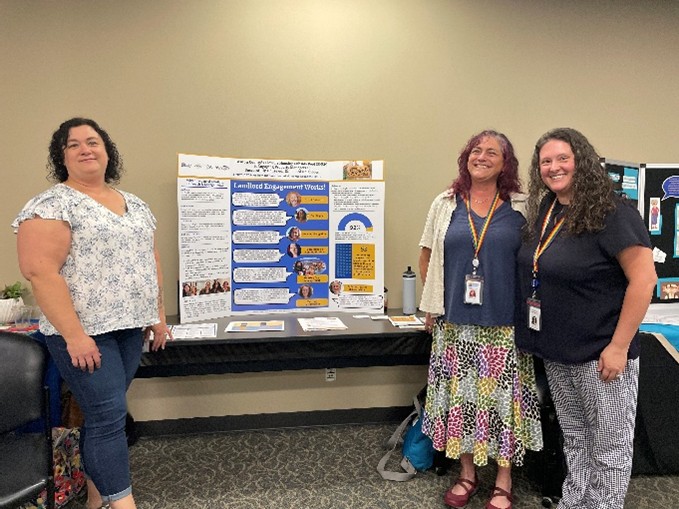 Three individuals stand beside a display table featuring an informational poster and printed materials at what appears to be an event or resource fair