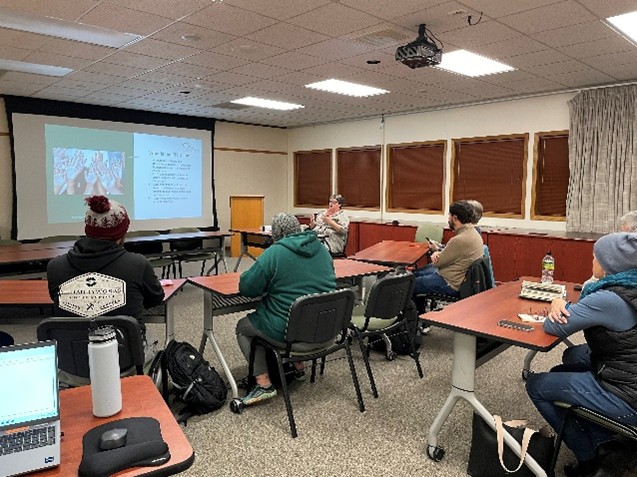 People seated in a classroom watch a presentation projected on a screen at the front of the room.