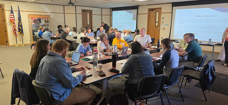 A group of people sitting in chairs and standing around tables in a conference room with projector screens displaying a PowerPoint presentation.