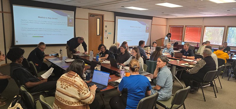 People working together around tables in a large room with laptops, worksheet packets, and notepads.