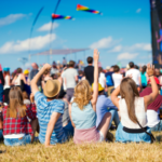 Young people sitting on the grass in a crowd during an outdoor music festival with rainbow flags in the distance.