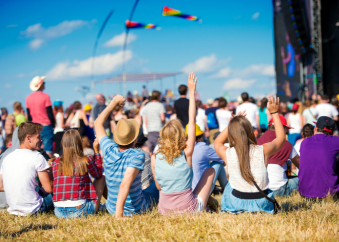 Young people sitting on the grass in a crowd during an outdoor music festival with rainbow flags in the distance.