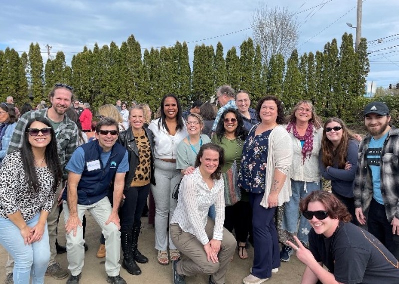 Group of people standing outdoors in front of tall greenery, gathered closely and looking toward the camera.