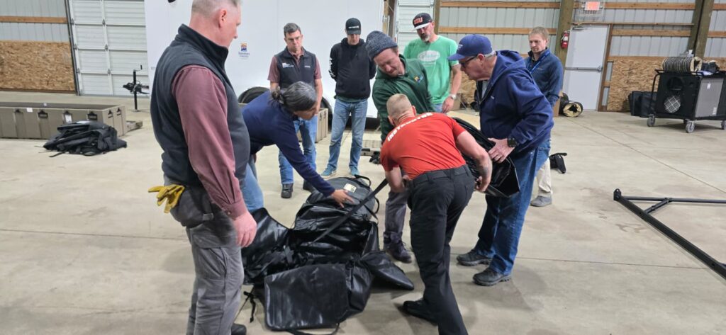 A group of people gathered in a warehouse working together to pack an emergency tent into a large black tarp on the floor.