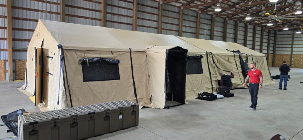 Large beige emergency tent set up inside a spacious warehouse with two men nearby and equipment cases on the floor.