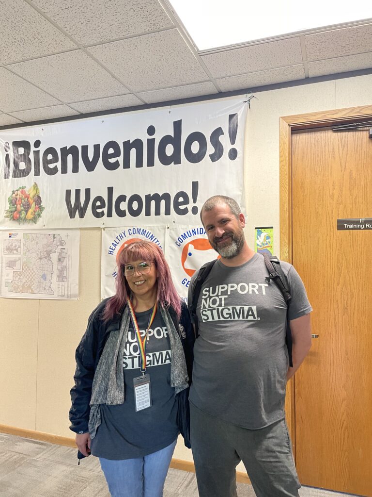 Two people wearing gray shirts that say, "Support Not Stigma," standing under a welcome banner.