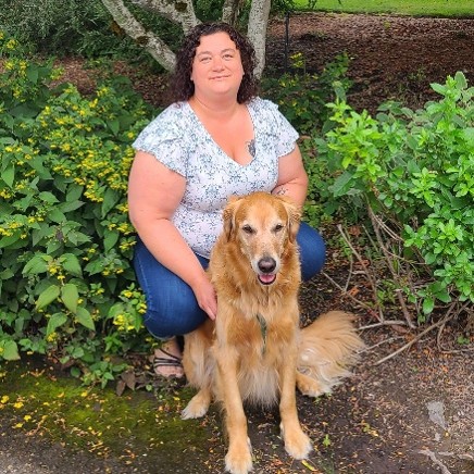A woman with curly brown hair wearing a floral top and jeans crouches down near some shrubbery with a sitting golden retriever.