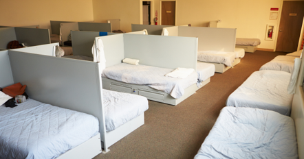 A shared dormitory-style sleeping space like in an emergency shelter with white beds and divider walls.