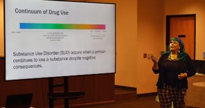 Presenter with blue hair explains a slide showing the continuum of drug use from no use to substance use disorder on a large screen.