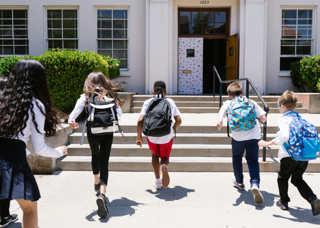 Five children with backpacks eagerly run up steps toward a school building entrance on a sunny day.