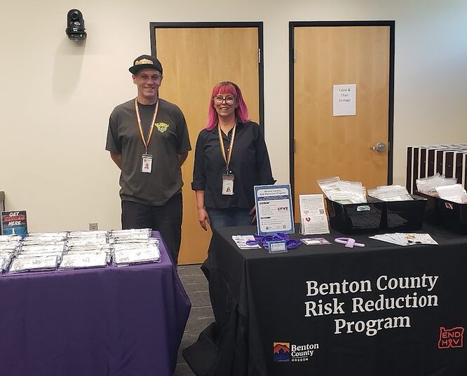 Two staff members stand behind tables displaying materials for the Benton County Risk Reduction Program in an indoor setting.
