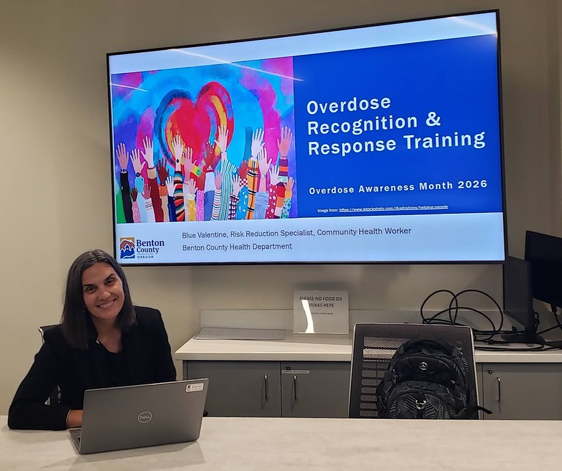 Woman sitting at a conference table with a laptop, next to a screen displaying 'Overdose Recognition & Response Training' for Overdose Awareness Month 2026.