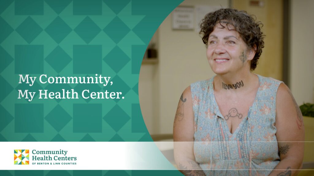Woman with short curly brown hair, covered in tattoos and wearing a sleeveless floral blouse promotes the “My Community My Health Centers” campaign.