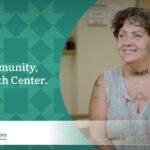 Woman with short curly brown hair, covered in tattoos and wearing a sleeveless floral blouse promotes the “My Community My Health Centers” campaign.