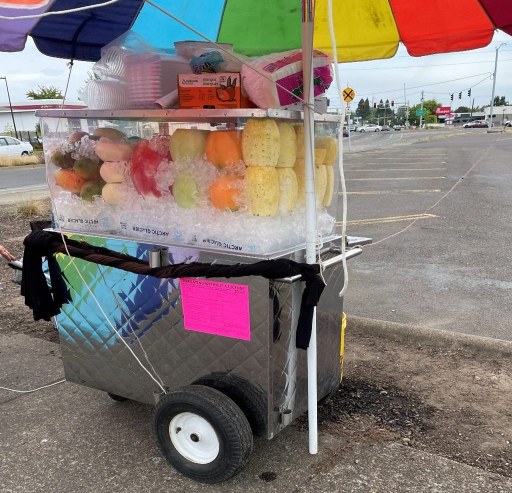 A cart selling cut fruit under a rainbow umbrella. A neon pink sticker declares it is "Operating Without a License."