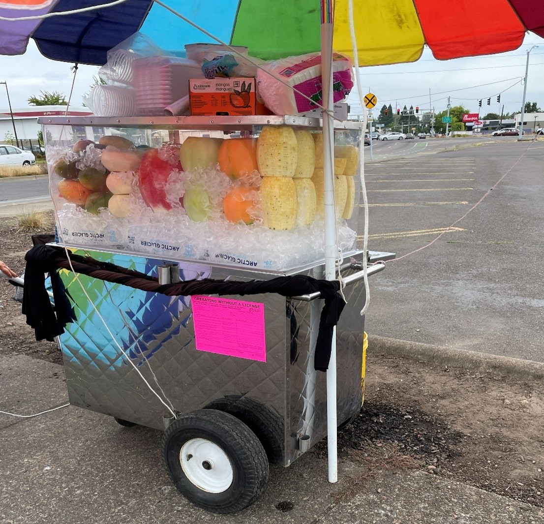 A cart selling cut fruit under a rainbow umbrella. A neon pink sticker declares it is "Operating Without a License."