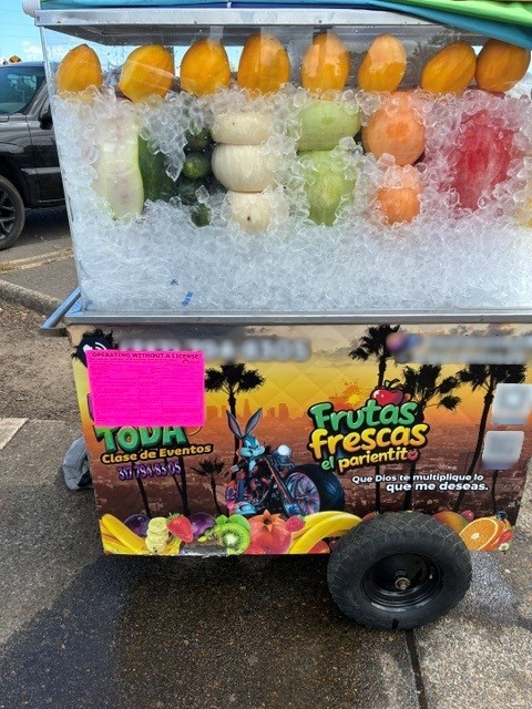 Unlicensed vendor selling cut fruit. A neon pink sticker is attached to their cart that says "Operating Without a License"