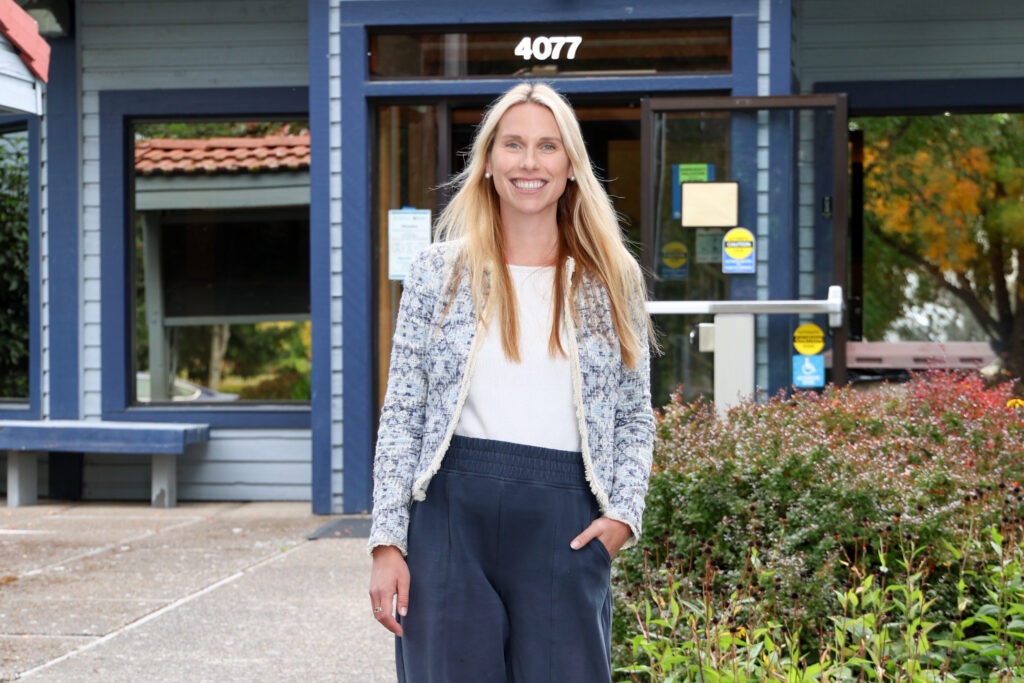 A person with long light-colored hair stands outdoors in front of a blue building entrance marked with the number 4077. There are plants and shrubs on one side of the walkway and large windows and an open glass door behind them.