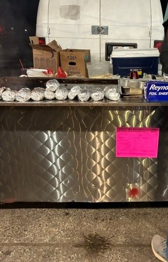 An unlicensed vendor's counter, with hot food items on it, wrapped in tin foil. There is a white van parked behind it with a cooler, various food service items in boxes, and no visible way to keep hot food at a safe temperature.