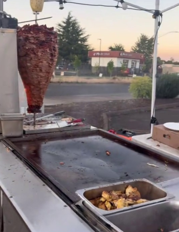 A large flat-top grill and vertical spit full of roasting meat, sitting in the corner of a parking lot next to an intersection. There are unused utensils and food scraps on the flat surfaces, and a table with boxes on it set up behind.